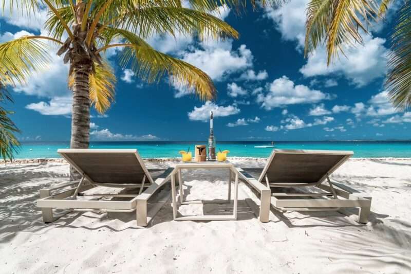 Two sun loungers under swaying palm trees on a white-sand beach at Delfins Beach Resort, Bonaire, overlooking the turquoise Caribbean Sea with tropical cocktails on the table